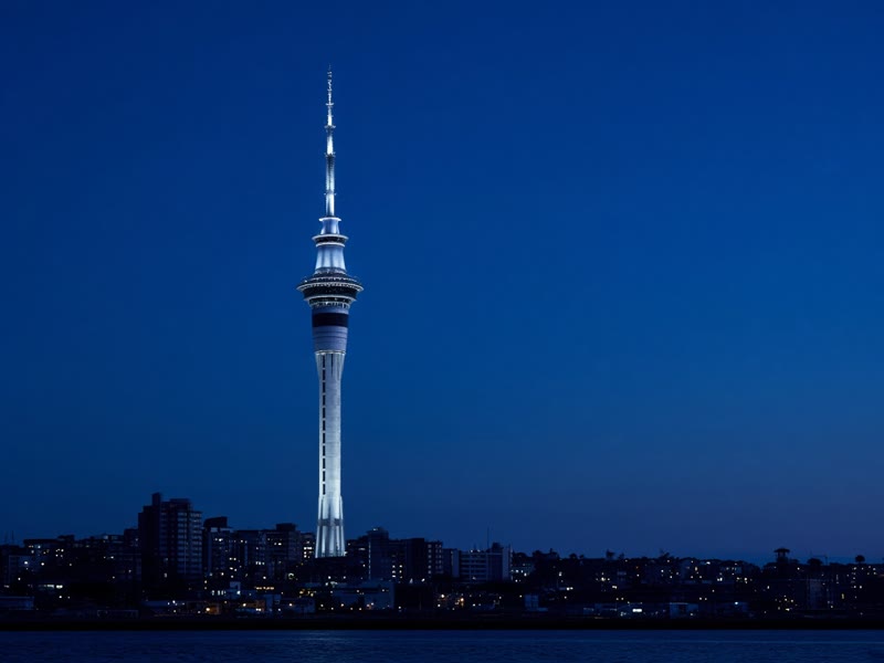 The Sky Tower lit against a blue evening sky above Auckland's harbour.
